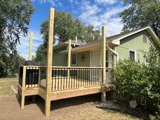 Wooden deck attached to a house with black railing and grill area, green house.