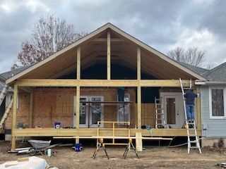 Construction of an outdoor deck and roof extension attached to a light blue house. Two workers are present.