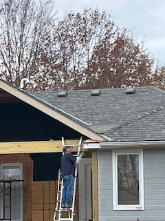 Person on a ladder, working on a house roof. Gray shingles, cloudy sky, trees in the background.