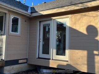 Exterior of house with new door and window, tan siding, white trim, shadow on wall, sunny day.