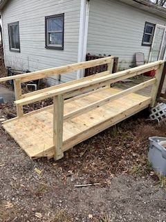 Wooden wheelchair ramp leading to a light green house with windows. Handrails on both sides.