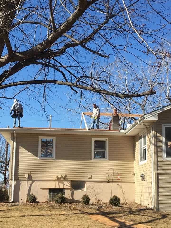 Two people on a beige house roof, working on a wooden frame, under a blue sky.