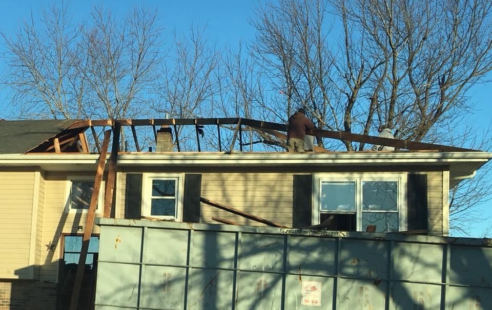 Person working on a partially demolished roof, above a dumpster, with bare trees and a blue sky.
