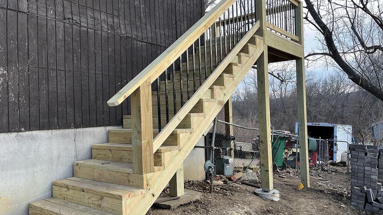 Wooden outdoor staircase attached to a dark wall and landing, featuring black metal railings.