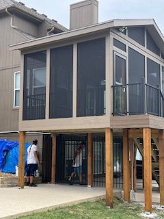 Screened porch on a raised deck, brown framing, two people painting beneath, grassy yard.