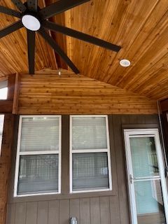 A porch with a wooden ceiling, fan, and windows with blinds. A door is on the right.
