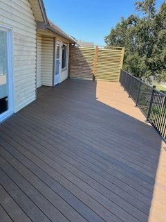 Wooden deck with a house on the left, a fence, and metal railing. Blue sky, brown deck, and green trees.