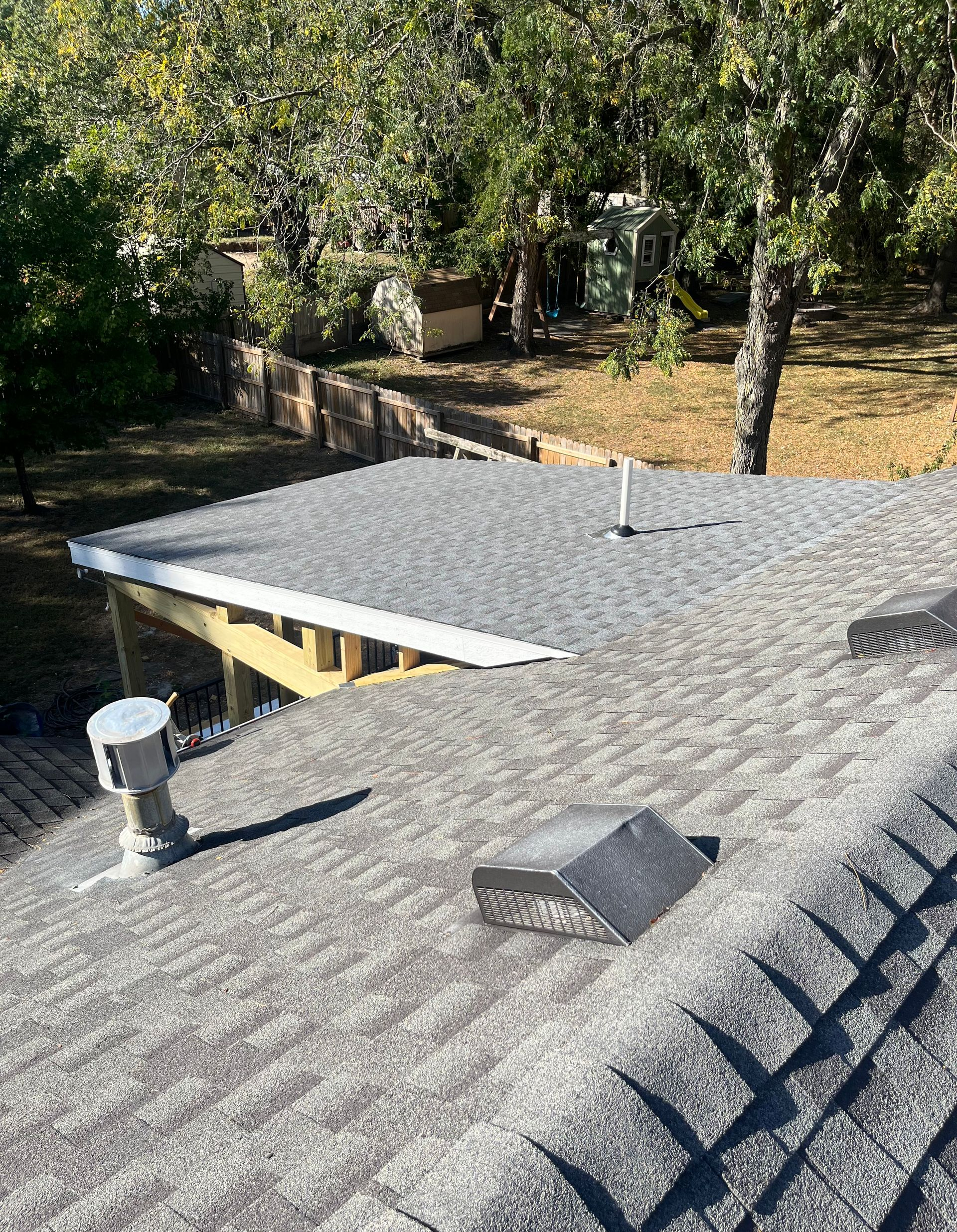 View of a gray shingled roof with a vent and a flat section. Trees and a yard are in the background.