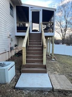 A screened porch with wooden stairs leading up from a concrete slab on a grassy yard.