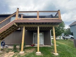 Elevated wooden deck with stairs and railing, supported by posts. Brown and tan colors. Overlooking a yard.