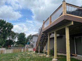 Wooden deck with stairs, supported by posts, against a house under a cloudy sky.