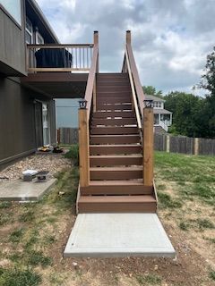 Wooden outdoor stairs lead up to a deck. Stairs and deck are brown. Concrete walkway at the base.