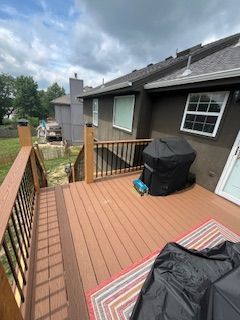 Wooden deck with brown and black railing, adjacent to a brown house, with a grill covered in black, and a rug.