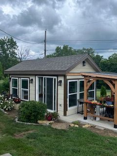 Small beige building with multiple windows, brown trim, and a pergola, set in a grassy yard under a cloudy sky.