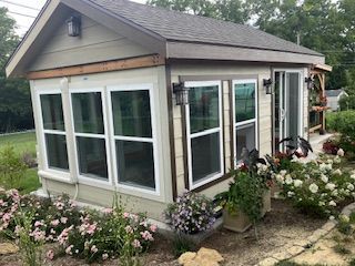 Small sunroom with white-framed windows and a sliding door. Beige siding, surrounded by a garden with pink flowers.
