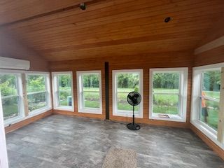 Sunroom with wood ceiling and paneling, gray floor, multiple windows, and a standing fan.