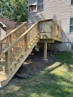Wooden deck and stairs leading up to a two-story house with green grass and dirt around the base.