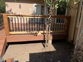 Wooden deck with black railing and bench, tree growing in dirt. House in background.