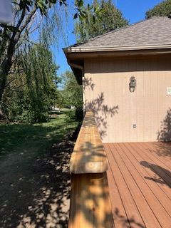 Wooden deck with railing next to a light-colored building with a brown roof and a grassy area with trees.