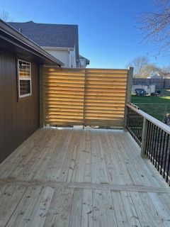Wooden deck with a horizontal slat privacy screen next to a house with black railings and a green yard.