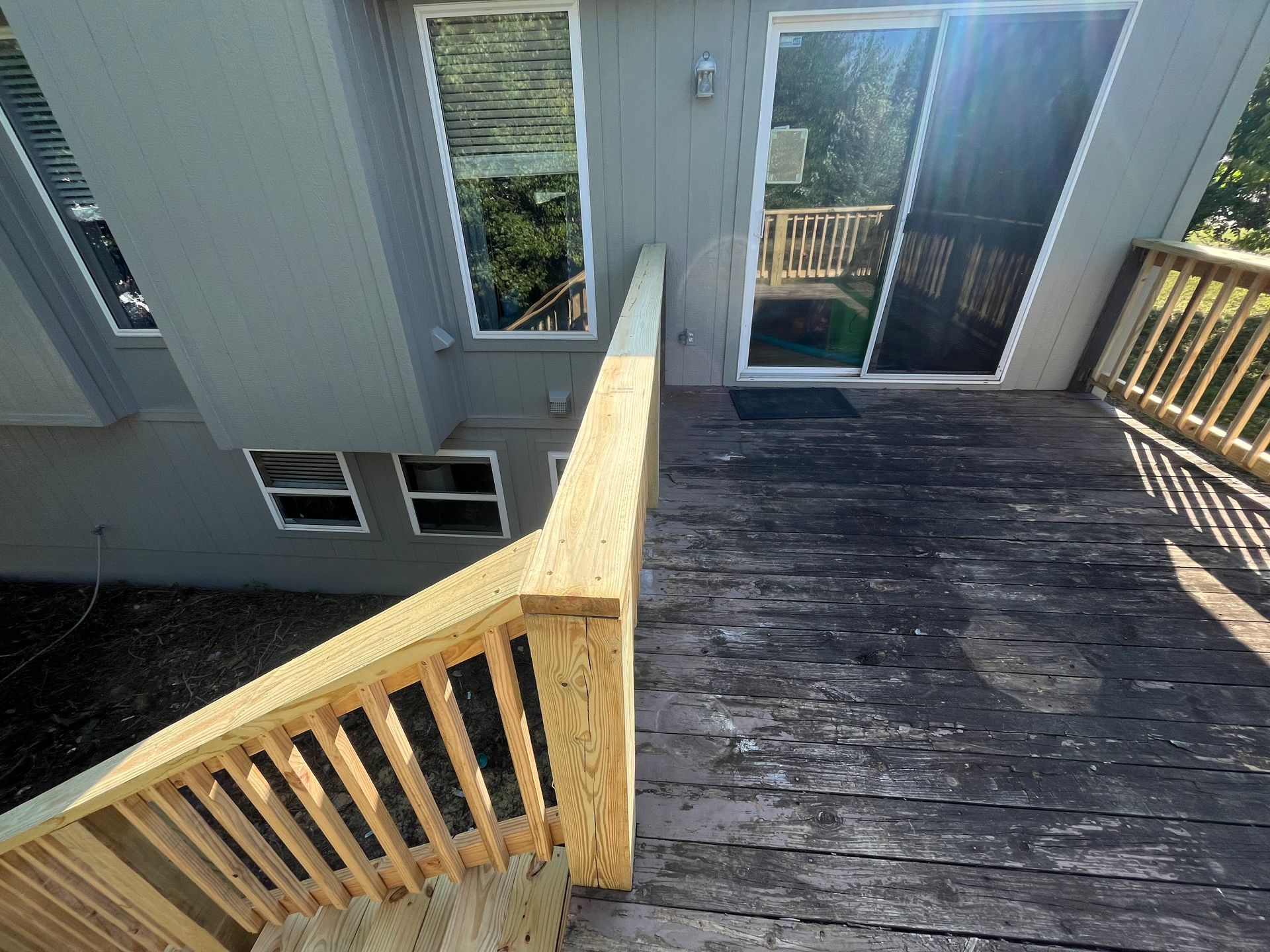 Exterior view of a deck with weathered boards and a new wooden railing. Sliding glass door and windows visible.