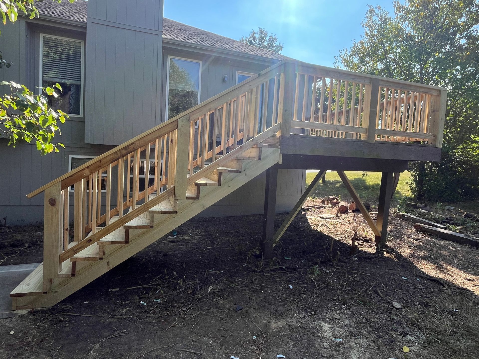 Wooden deck with stairs attached to a light-colored house. Brown deck boards and railings against a blue sky.