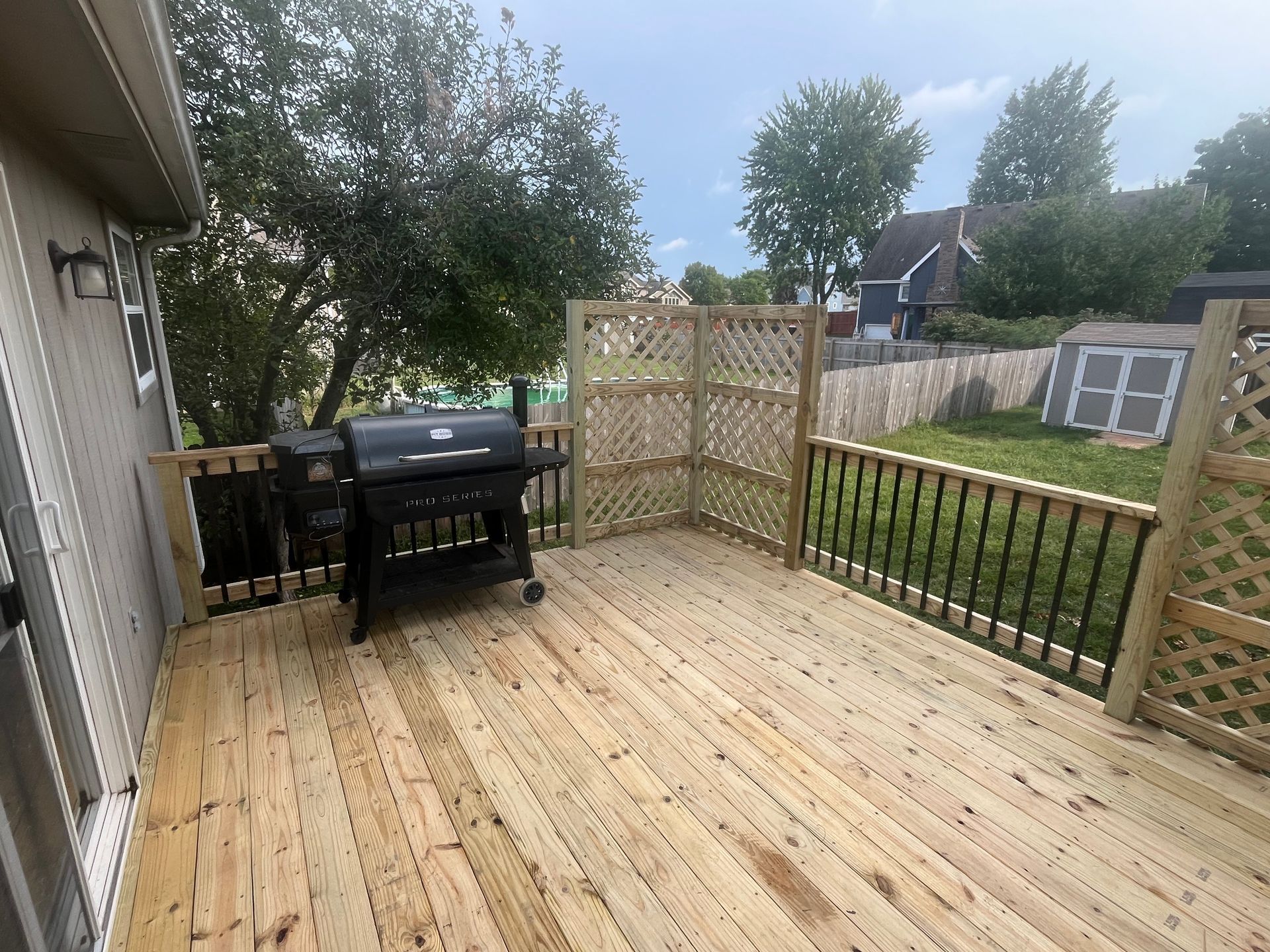 Wooden deck with black grill, lattice screens, and black railing. Backyard view with shed and fence.