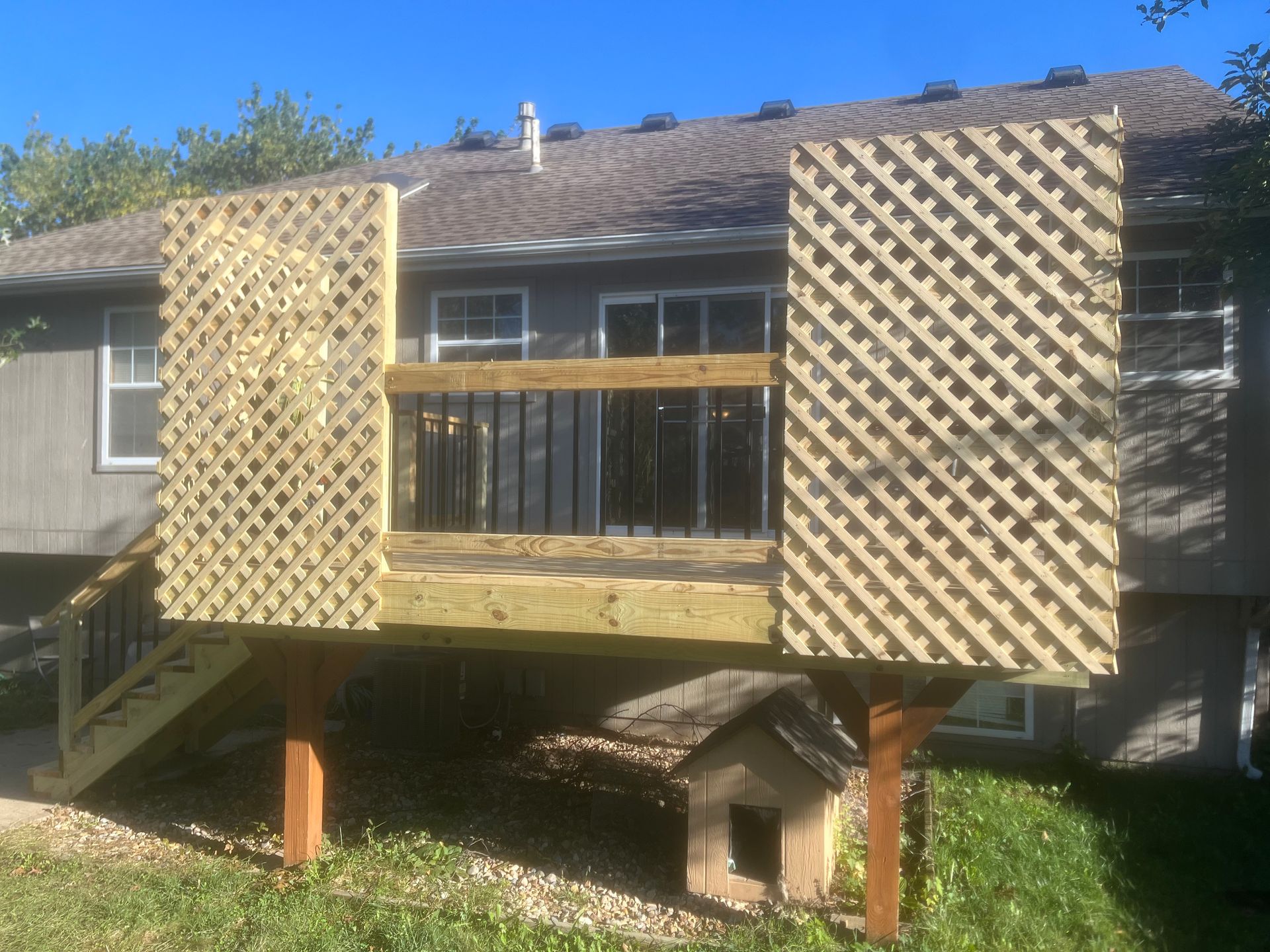 Wooden deck with lattice panels, steps, and a dog house in a yard.