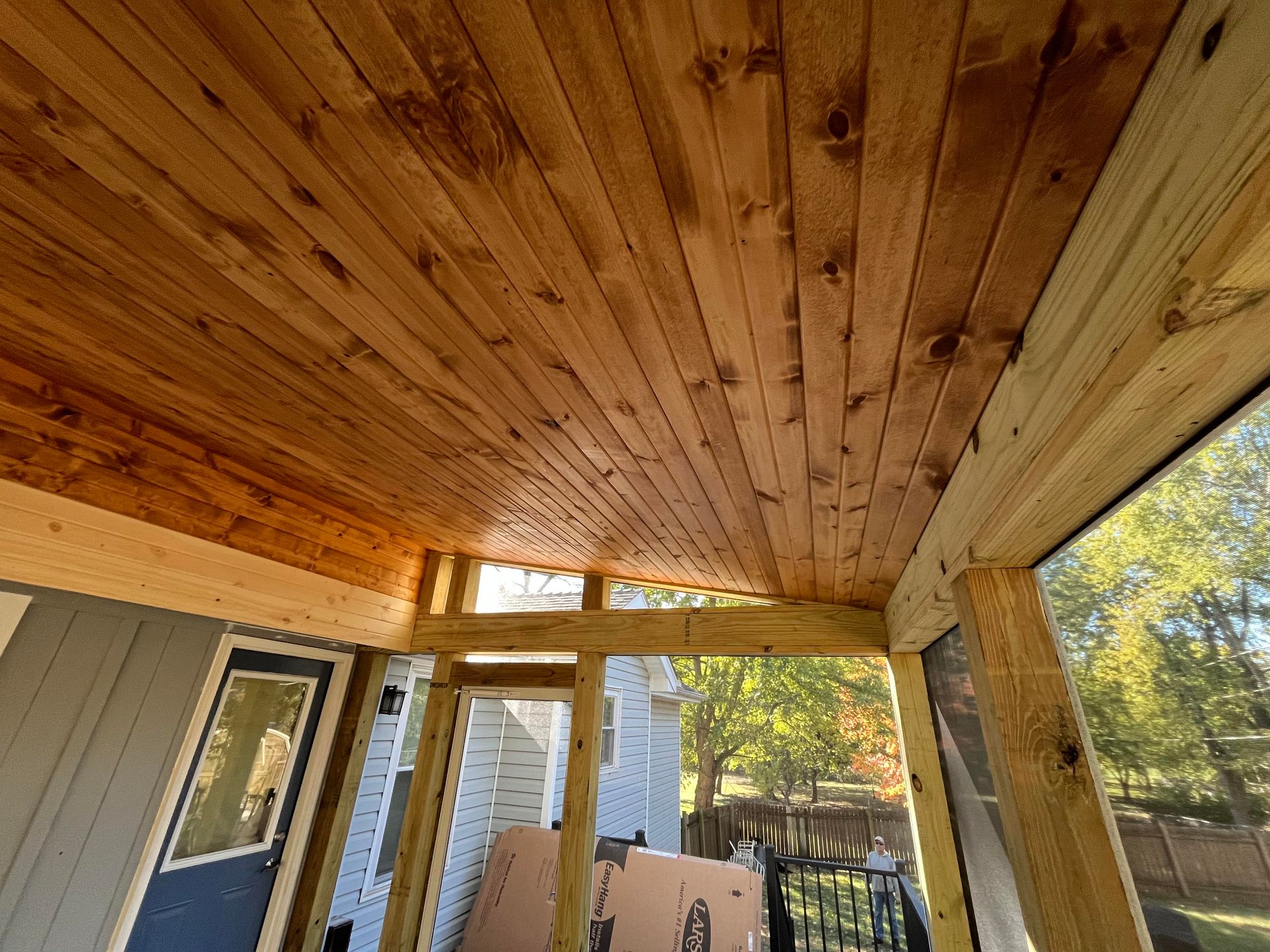 Wooden ceiling of a screened porch, stained brown, supported by light wood beams.