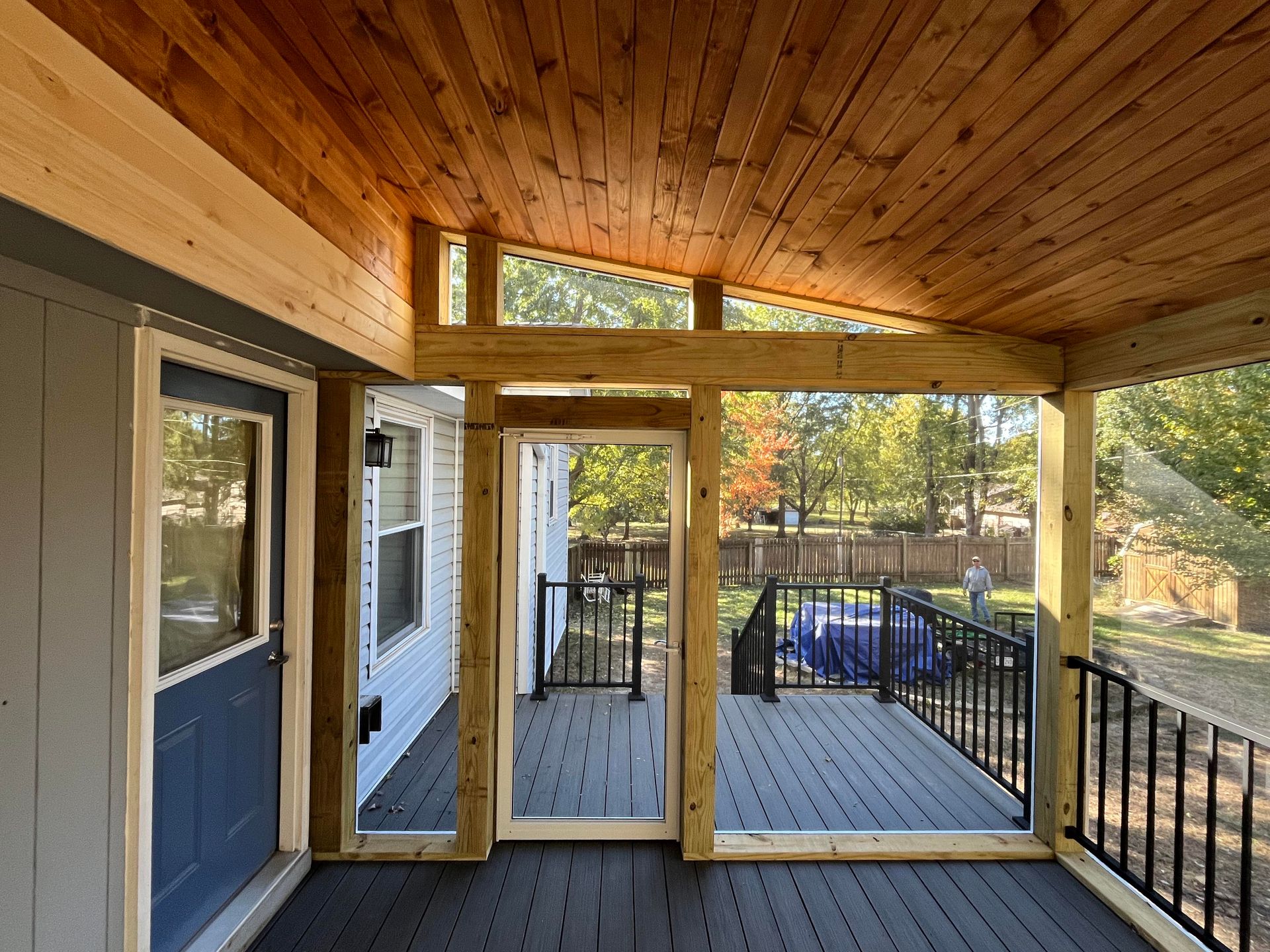 Covered porch with wood ceiling and frame, gray deck, blue door, and black railing.