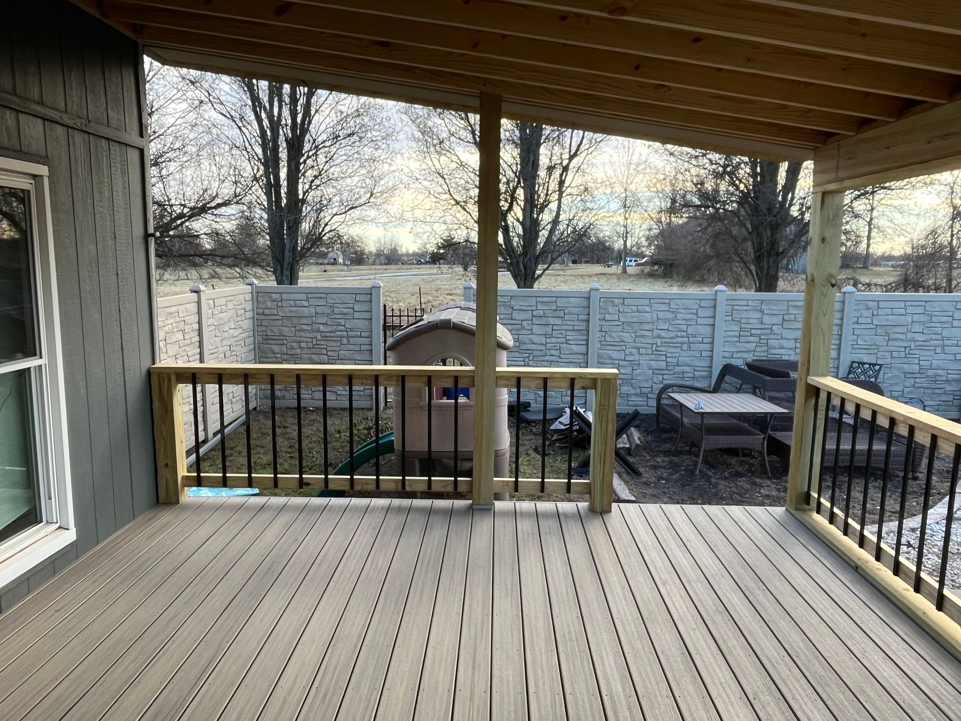 Covered deck with gray composite decking, black railing, and a view of a backyard with a playhouse and stone wall.