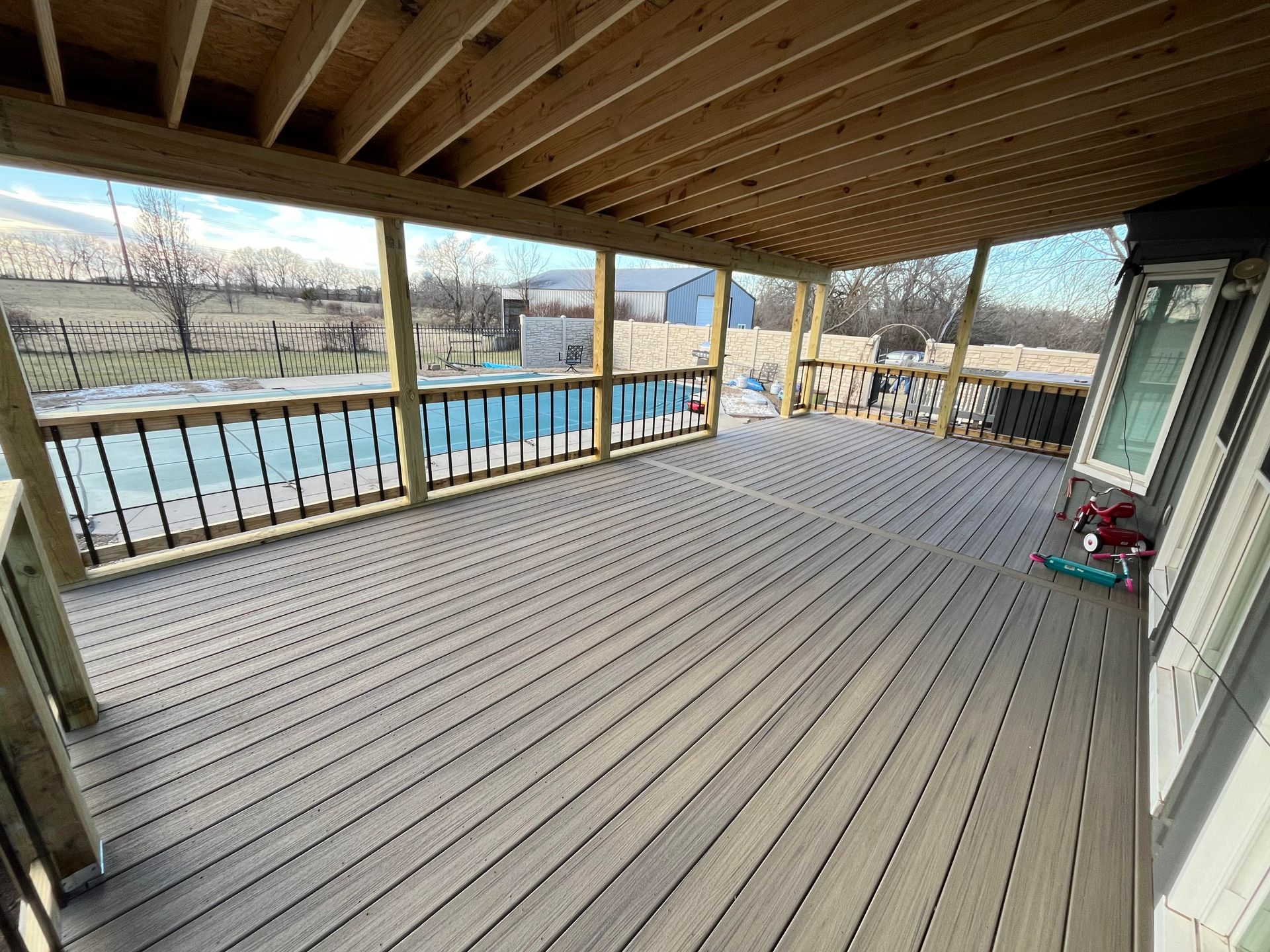 Covered wooden deck with black railing overlooking a pool and yard on a sunny day.