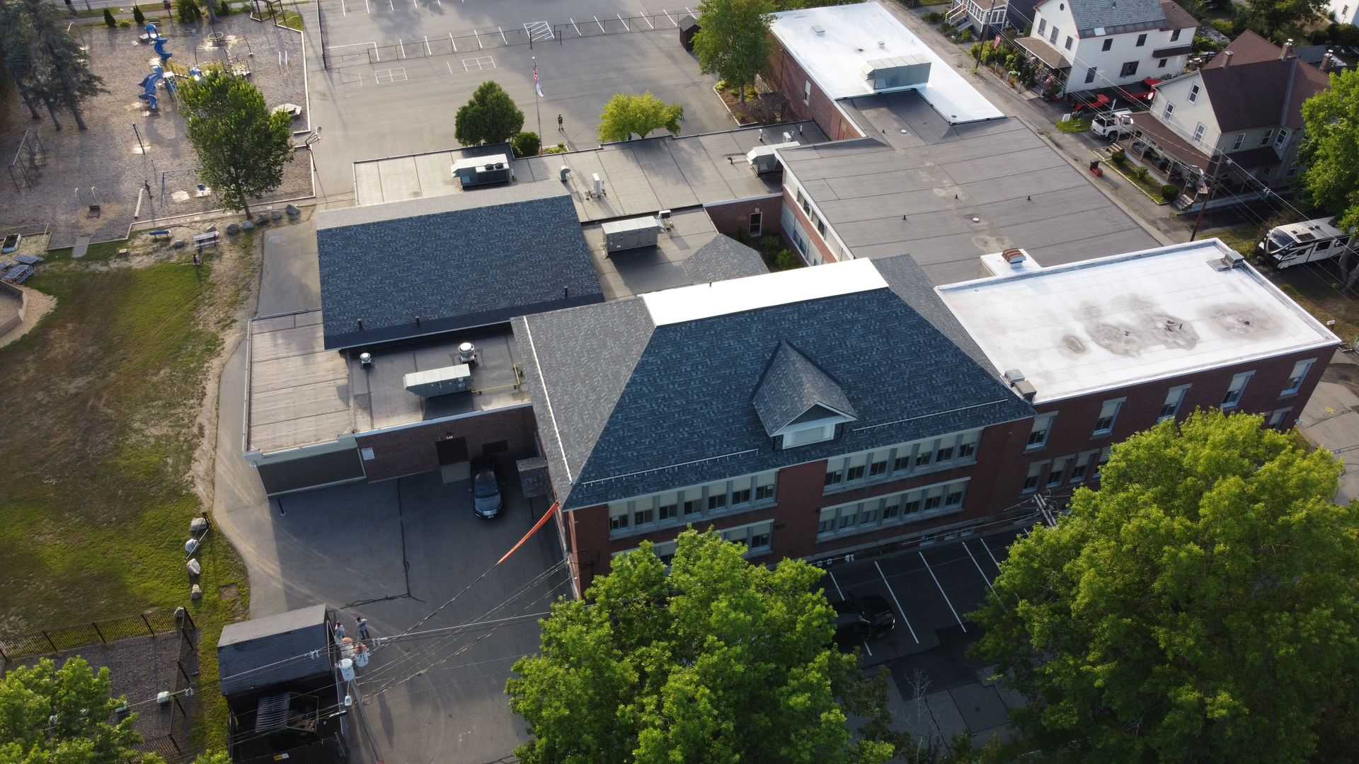 Aerial view of brick building with dark gray roof, trees, and paved areas.
