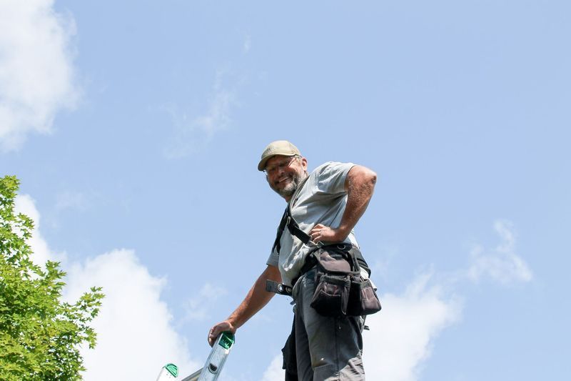 Roofer on ladder smiles, sunny day, blue sky, tool belt, holding ladder, green trees in background.