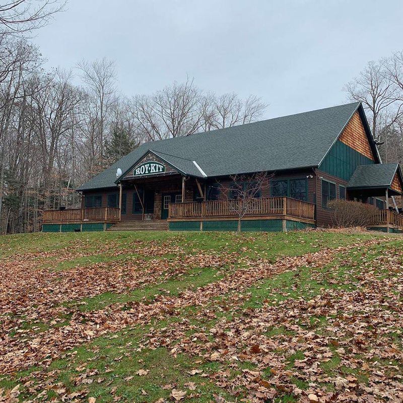 Brown building with a dark green roof and wooden deck, in a wooded area with a grassy hill covered in fall leaves.