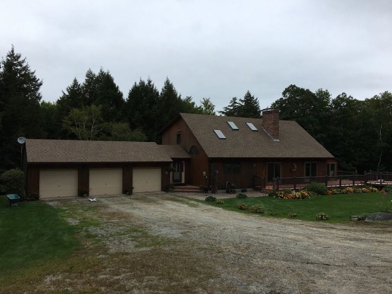 Brown house with attached three-car garage, gravel driveway, and surrounding trees under an overcast sky.