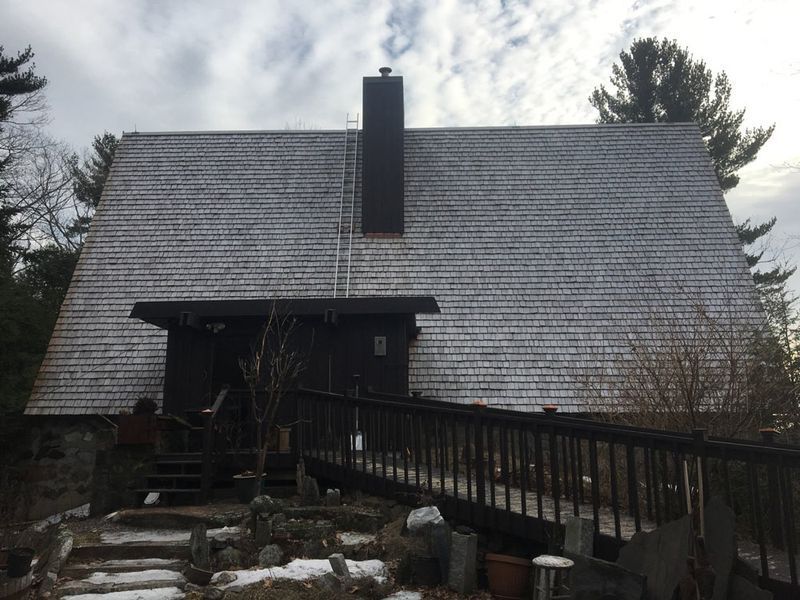 Wooden cabin with shake roof, snow, chimney, ramp, and stone steps. Overcast day.