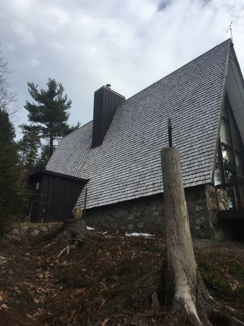 A-frame house with stone base and cedar shake roof, set on a hillside with a dead tree in the foreground.