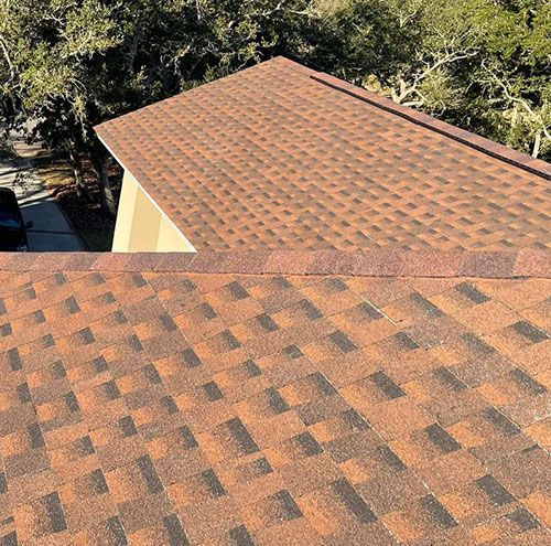 Two tan shingled rooftops seen from above, with trees in the background.