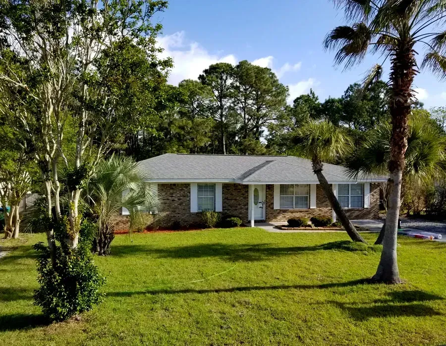 Single-story suburban house with a green lawn, palm trees, and a wooded backyard under a blue sky