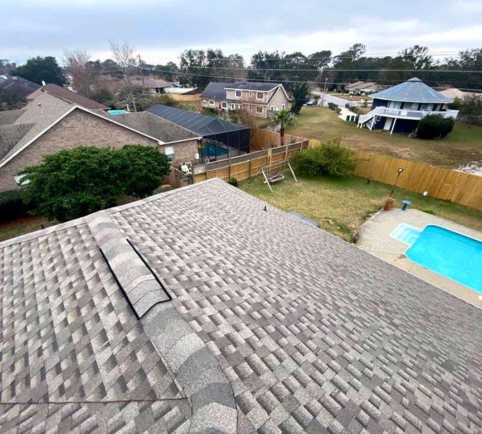 View from a shingled rooftop over suburban homes, fenced yard, and backyard swimming pool
