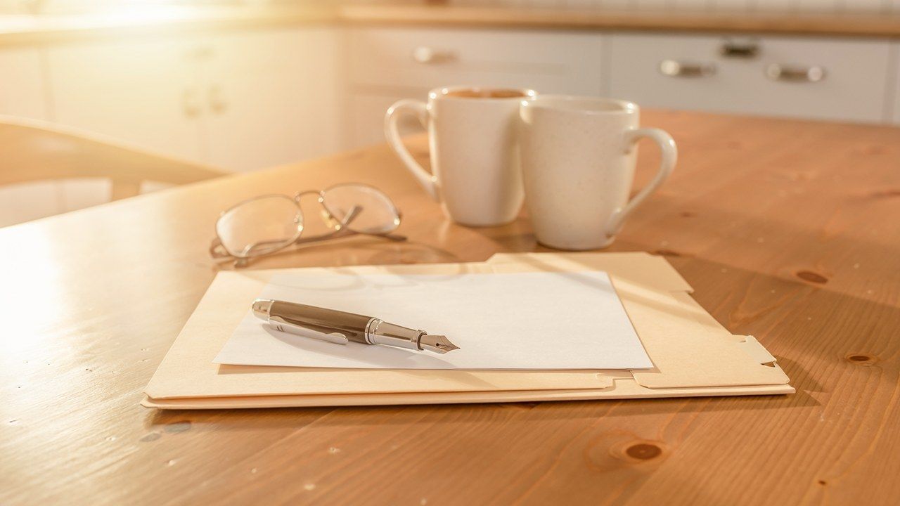 A manila folder, fountain pen, blank paper, reading glasses, and two coffee mugs on a sunlit wooden dining table.