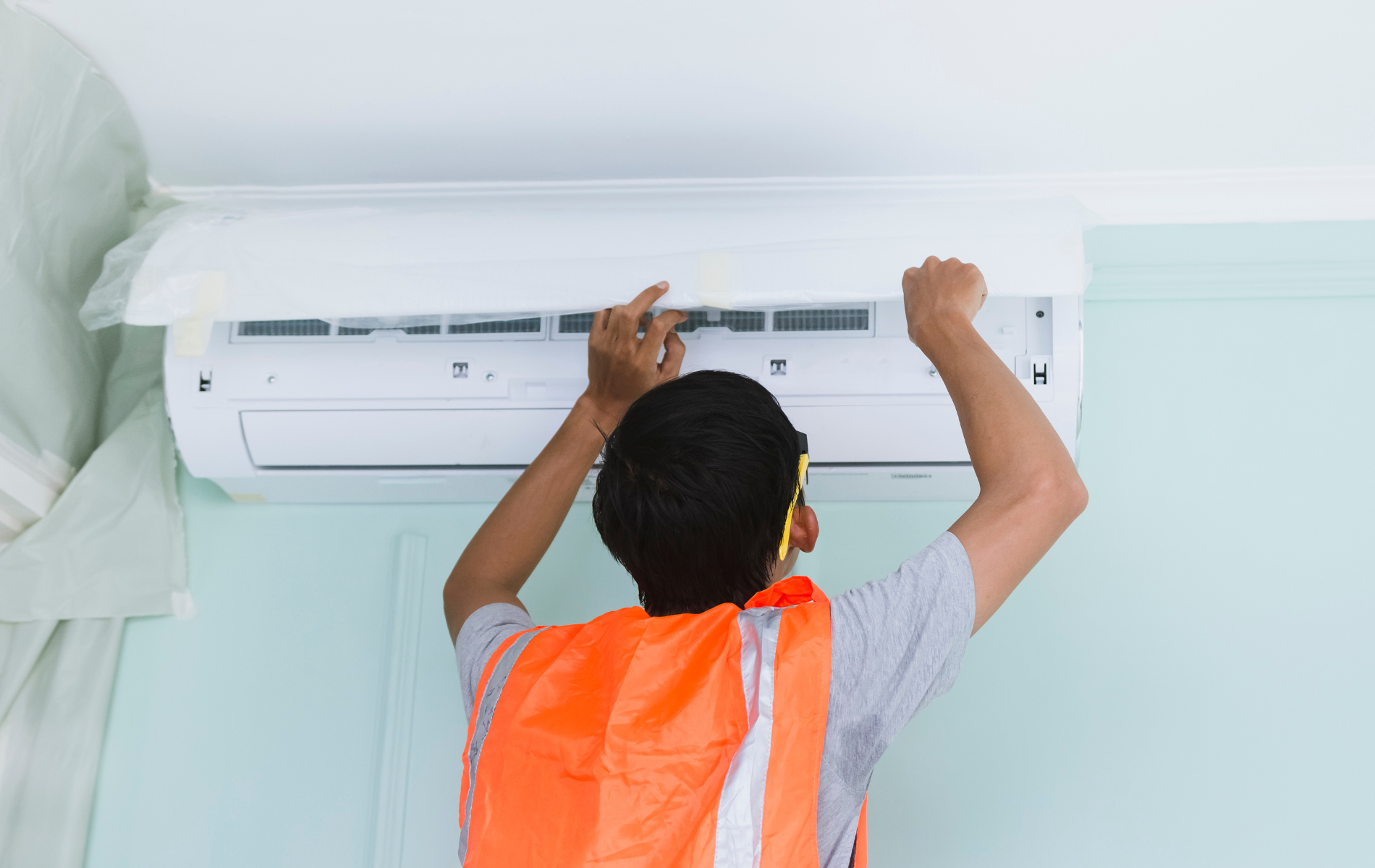 Person in orange vest repairing a wall-mounted air conditioning unit. Hands working on the white appliance. Light green wall.