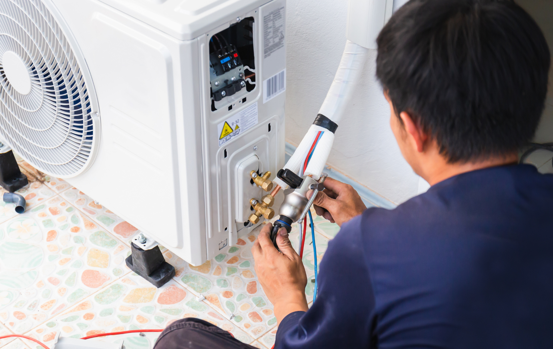 Person installing an air conditioner unit, connecting wires and pipes outdoors.