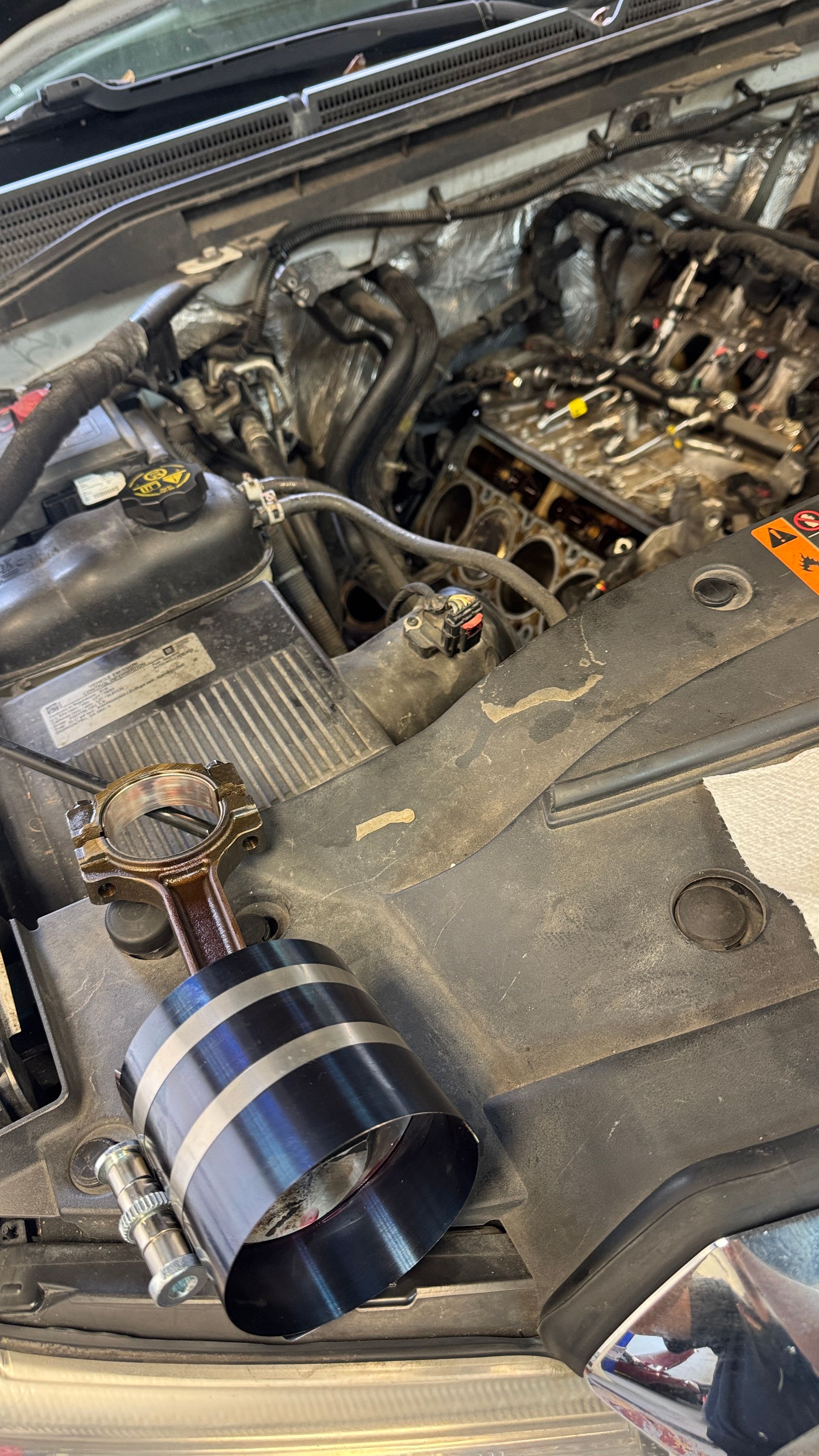 A piston with a black ring compressor installed sits on a car engine bay next to an exposed engine block.