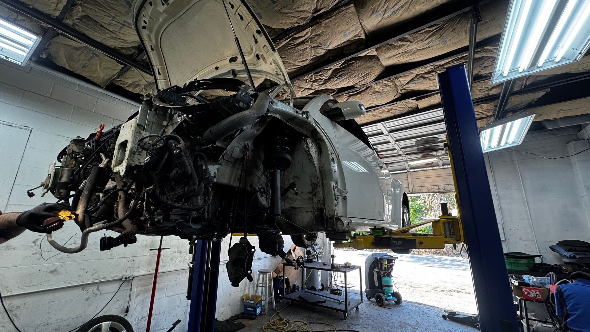A white car lifted on an auto repair shop lift, with its front bumper and engine components removed for maintenance.