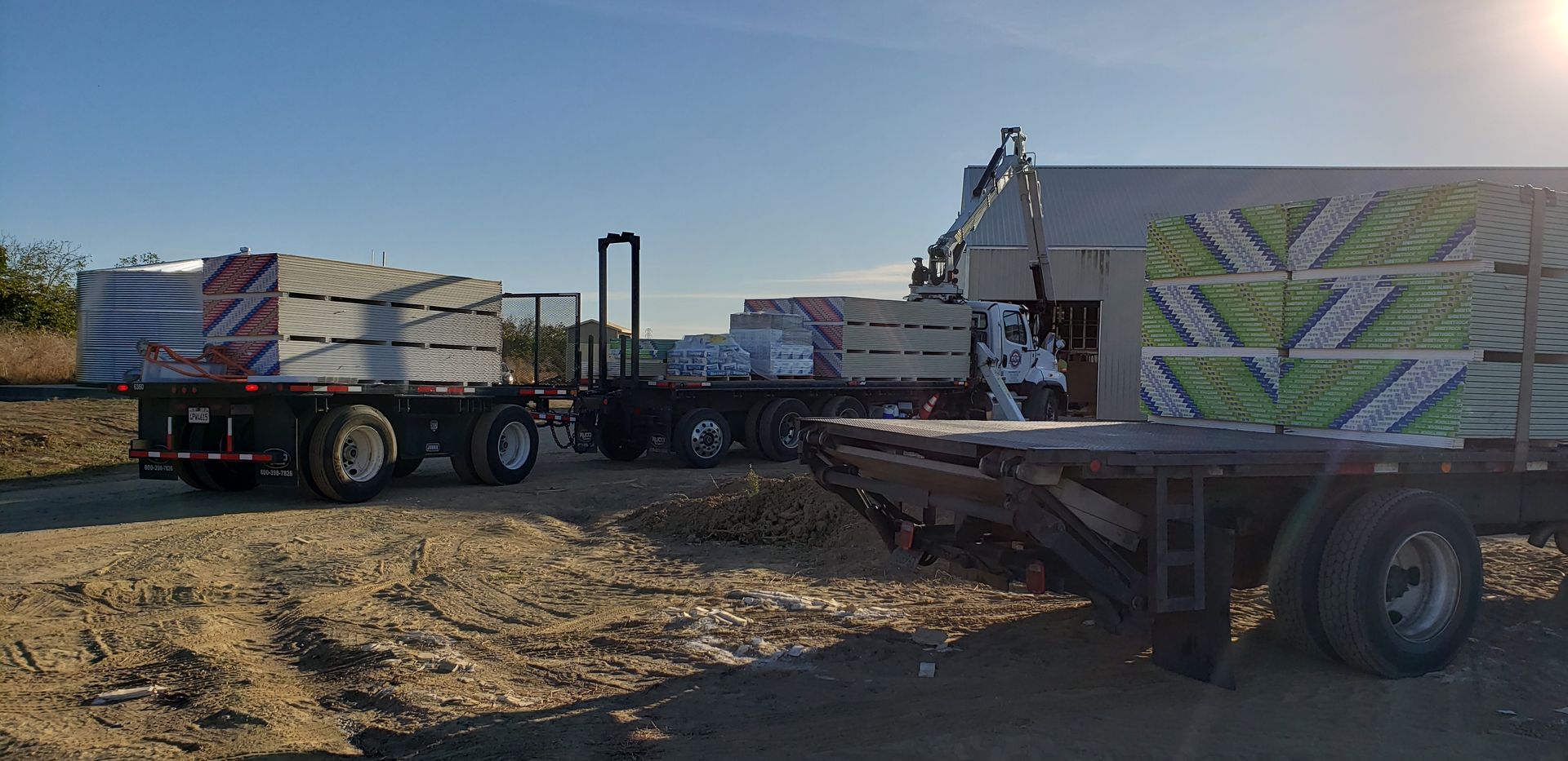 Flatbed trucks loaded with stacked materials at a dusty outdoor worksite under clear skies
