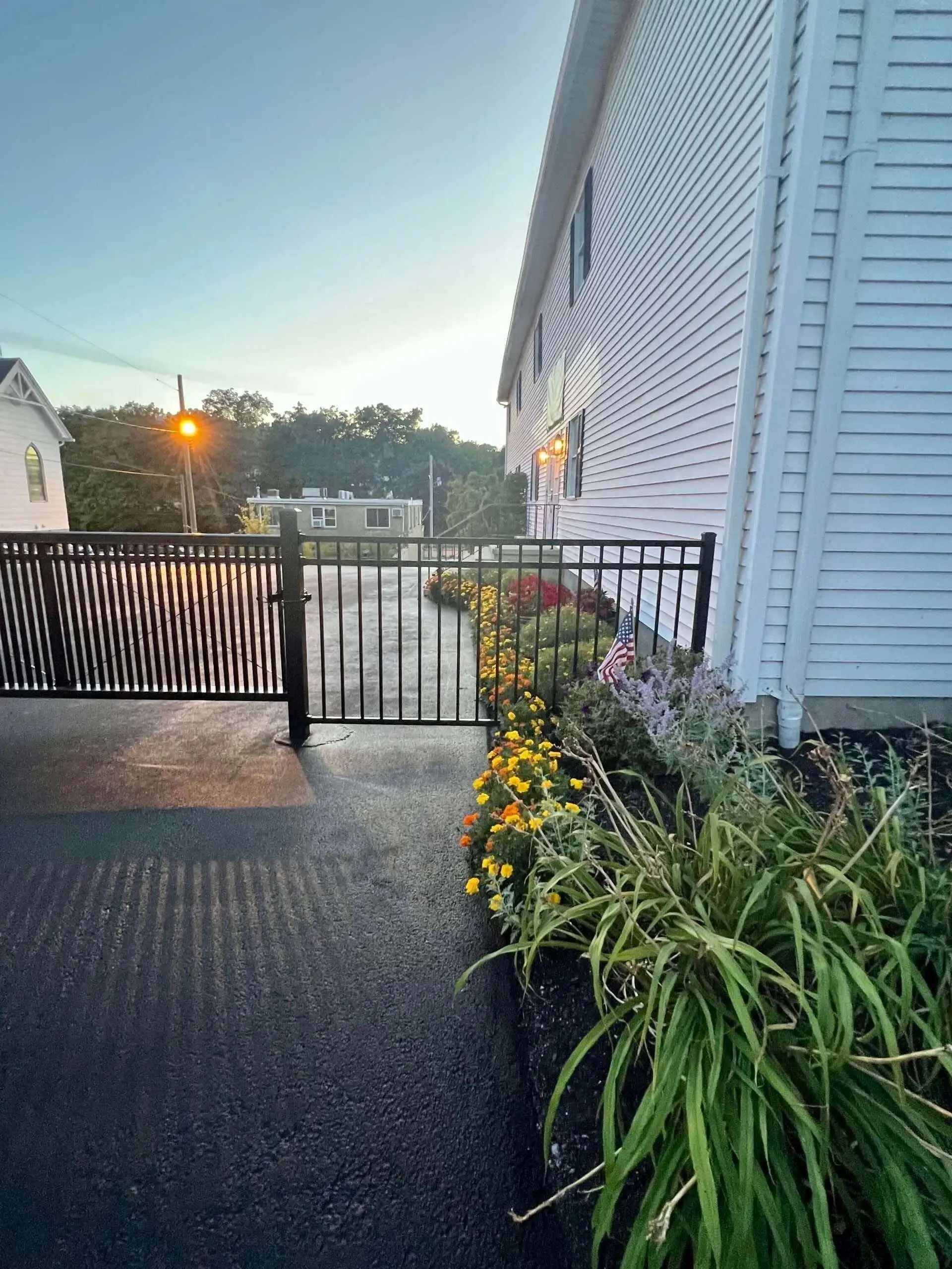 Black gate, flowers, and building. Paved area with street and trees in the distance.