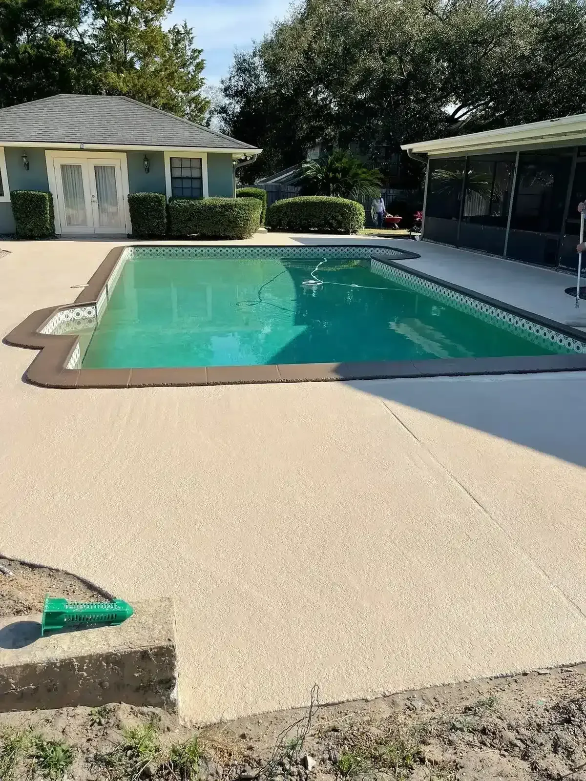 Backyard swimming pool beside a patio and small building, with turquoise water and surrounding trees.