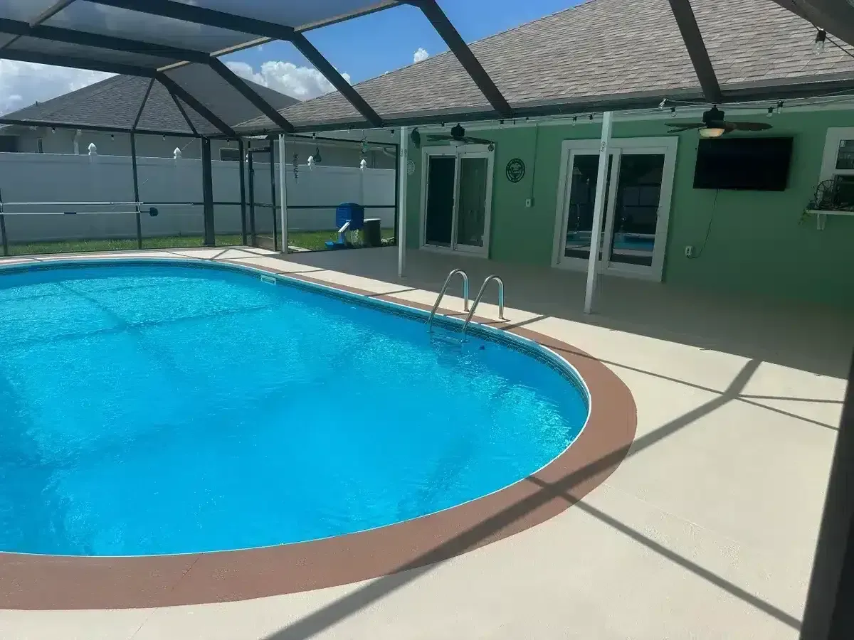 Covered backyard pool with bright blue water beside a green house and patio.